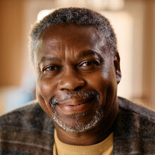 Older black man wearing a gold sweater and patterned vest smiles in his home.