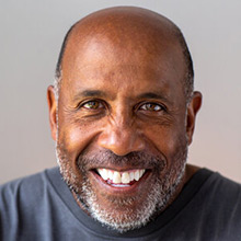 Older black man in pale blue t-shirt smiles.